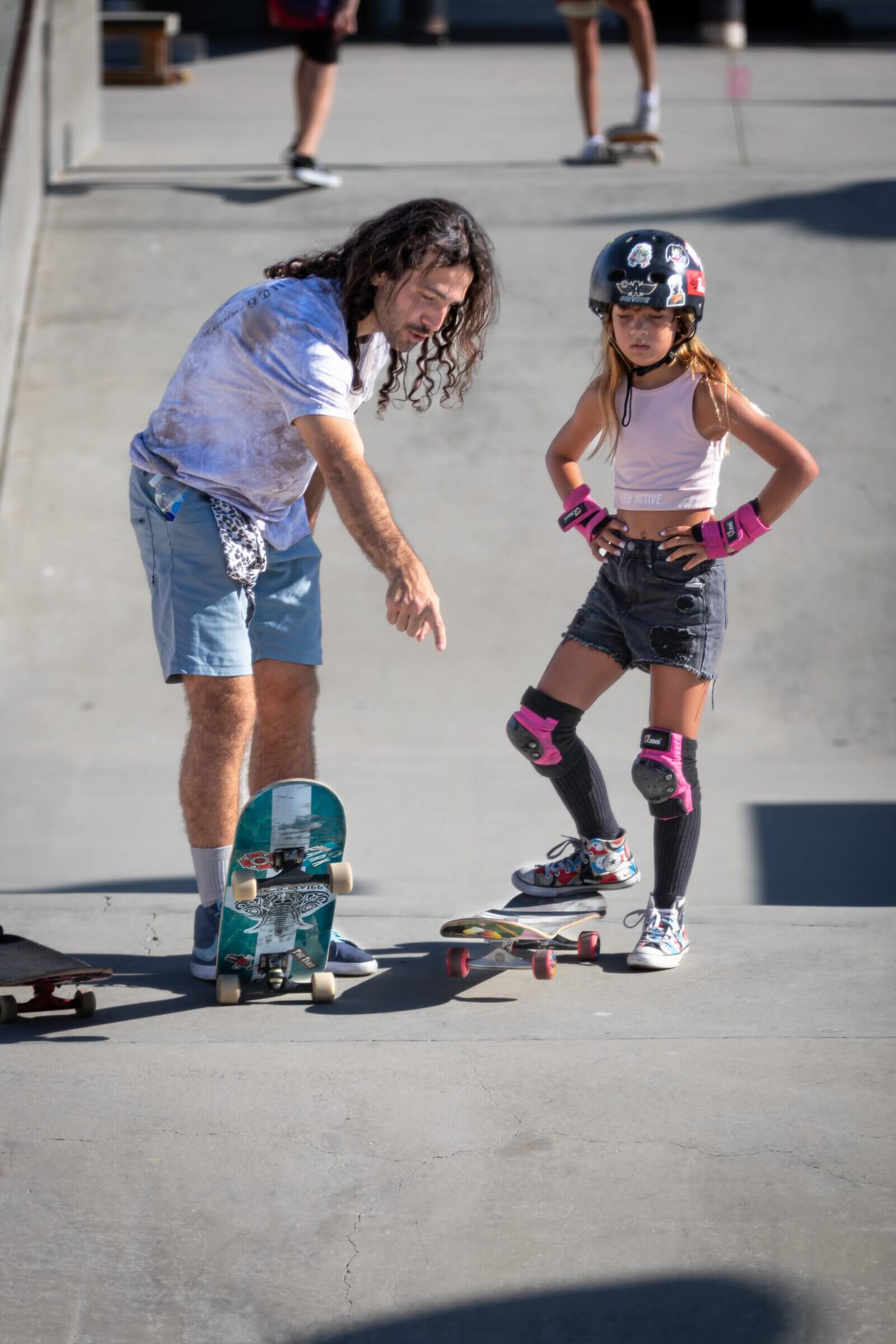 An instructor coaches a helmeted young skater at the skatepark.
